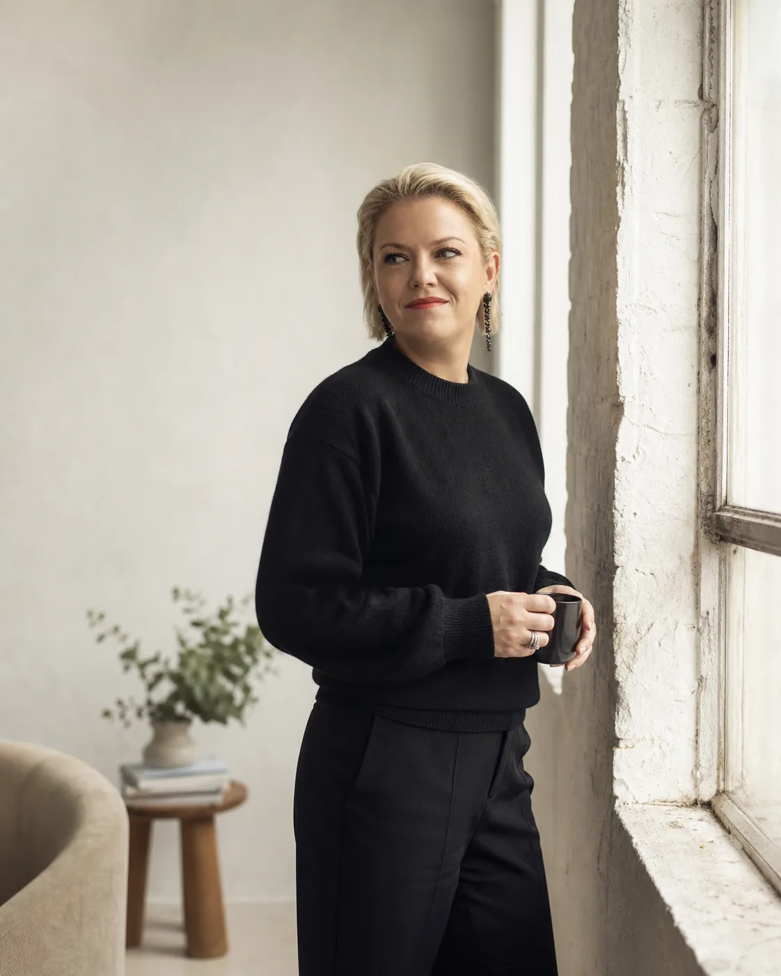 Michelle Sadlier by a tall window with a coffee, in a black knit