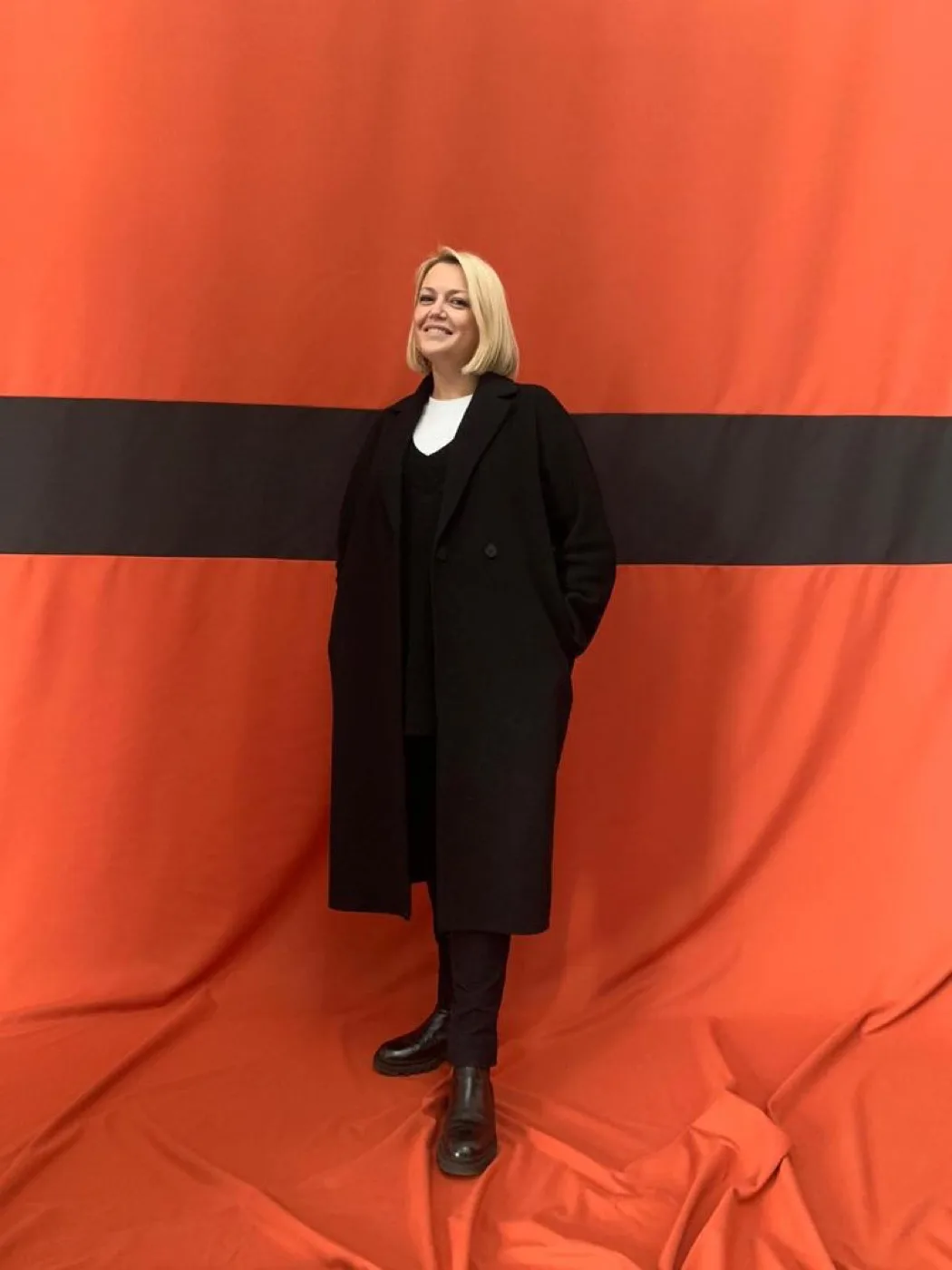 Michelle Sadlier in a long black coat against a vermilion show backdrop
